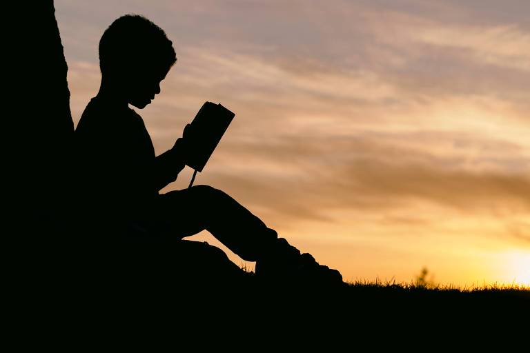 Foto mostra silhueta de criança sentada em um gramado e lendo um livro. No fundo, um céu amarelado indica o pôr do sol.