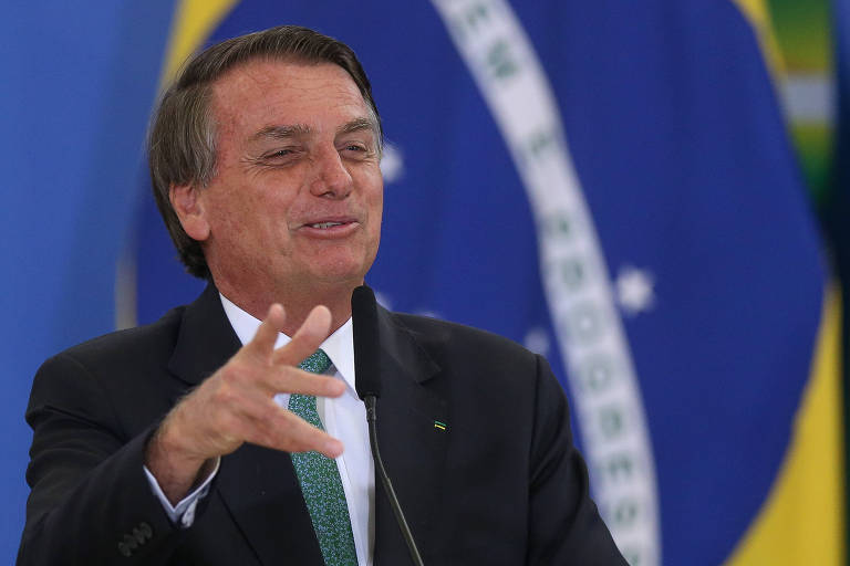 Bolsonaro aparece da barriga para cima. Ele é um homem branco de cabelos grisalhos, veste terno com gravata verde e gesticula com a mão direita em frente a uma grande bandeira do Brasil, discursando em evento.