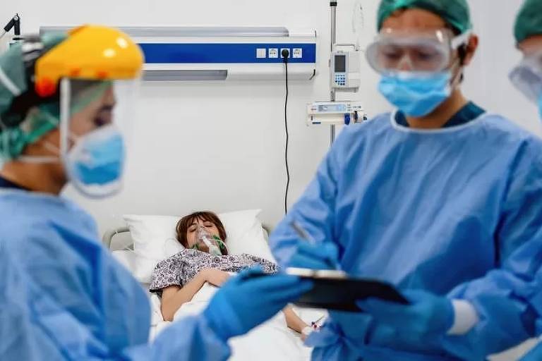 Em destaque, há três profissionais da saúde com roupa especial e máscara. Ao fundo, há uma paciente deitada em uma maca.