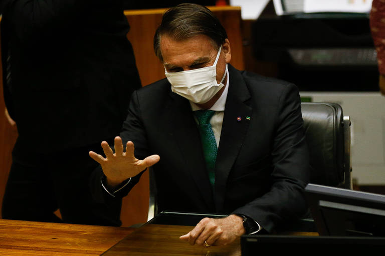 De máscara branca e sentado, Bolsonaro, um homem branco, cabelos castanhos e ternos, acena com uma das mãos aberta