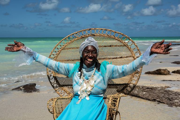 Em foto colorida, a cirandeira Lia de Itamaracá posa para a câmera sorrindo, de braços abertos e sentada em uma poltrona de vime em uma praia