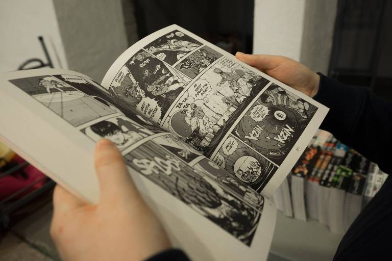 Foto colorida mostra mangá aberto, história em quadrinhos japonesa, segurado por duas mãos de uma pessoa 