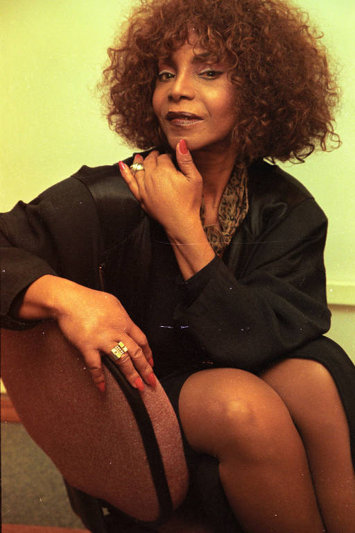 retrato colorido da cantora Elza Soares. ela está sentada de lado, apoiando-se no encosto de uma cadeira, posando para a foto 