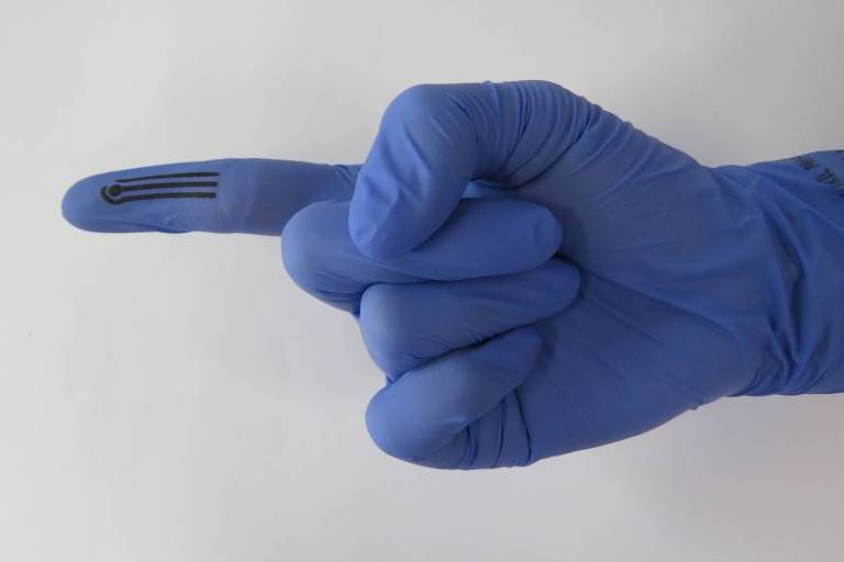 foto mostra mão usando luva azul de borracha; na ponta do dedo indicador, uma pequena superfície preta, onde estão os sensores do dispositivo 