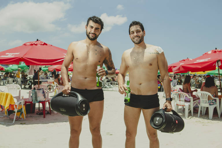 dois homens brancos em pé na faixa de areia da praia, em um dia ensolarado, cercados por guarda-sóis. os dois vestem sunga preta e cada um segura uma caixa de som 