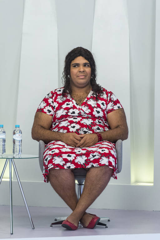 Paulo Vieira vestido como mulher, com vestido e peruca, sentado com as mãos no colo