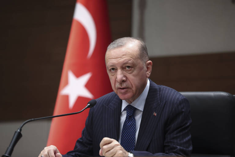 O presidente da Turquia, homem careca e de bigode, é visto sentado, durante um discurso, com um pequeno microfone em sua frente; atrás de si está a bandeira da Turquia
