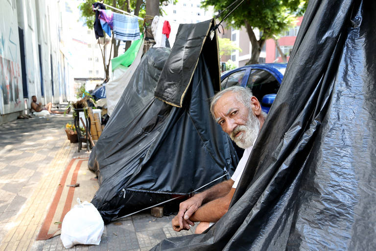 Homem idoso em situação de rua sentado ao lado de uma baraca feita de plástico reto