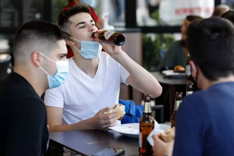 Imagem em primeiro plano mostra três pessoas sentadas em uma mesa de bar. Uma delas está com a máscara abaixada e leva uma garrafa a boca