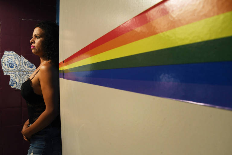 Mulher trans, encostada no lado esquerde uma parede, com as cores do arco-íris pintadas na parede bege à direita, como se estivessem saindo de suas costas.
