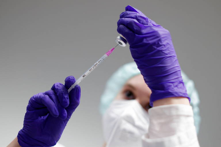 Enfermeira segura seringa com as mãos, protegidas com luvas na cor roxa, uma seringa e um frasco de vacina contra a Covid-19 da Pfizer
