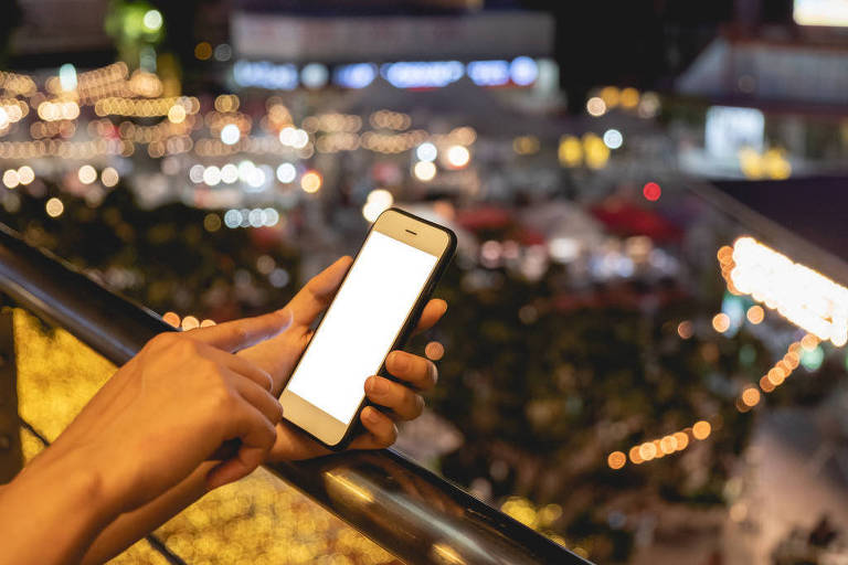 Pessoa usa o celular, com imagem de uma cidade ao fundo 