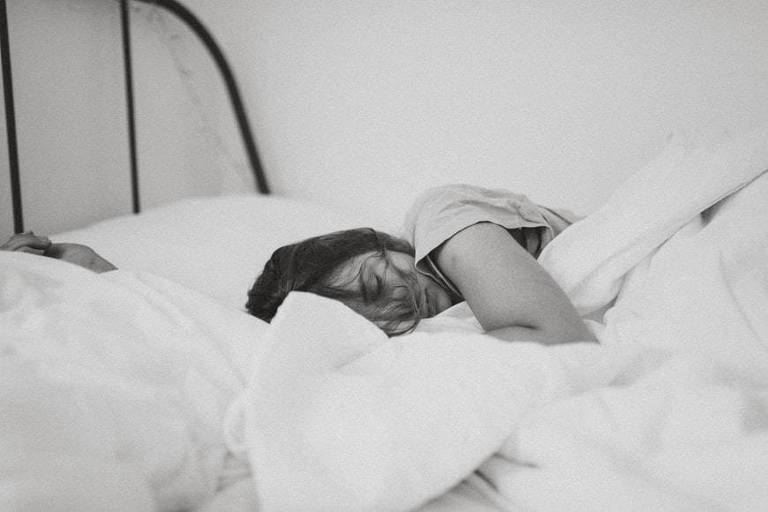 Foto em preto e branco mostra mulher dormindo em cama com travesseiros e lençóis brancos