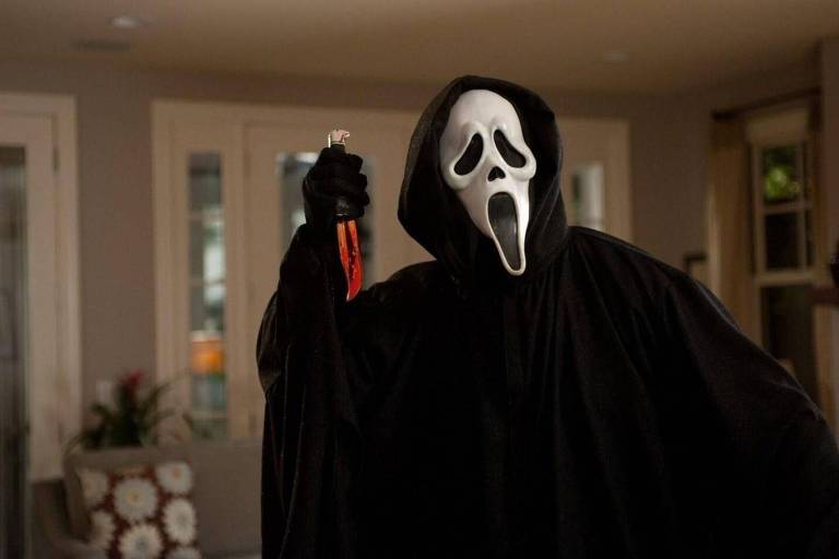 Foto colorida mostra frame do filme Pânico. Uma pessoa disfarçada com uma túnica preta e uma máscara de terror branca segura uma faca ensanguentada.