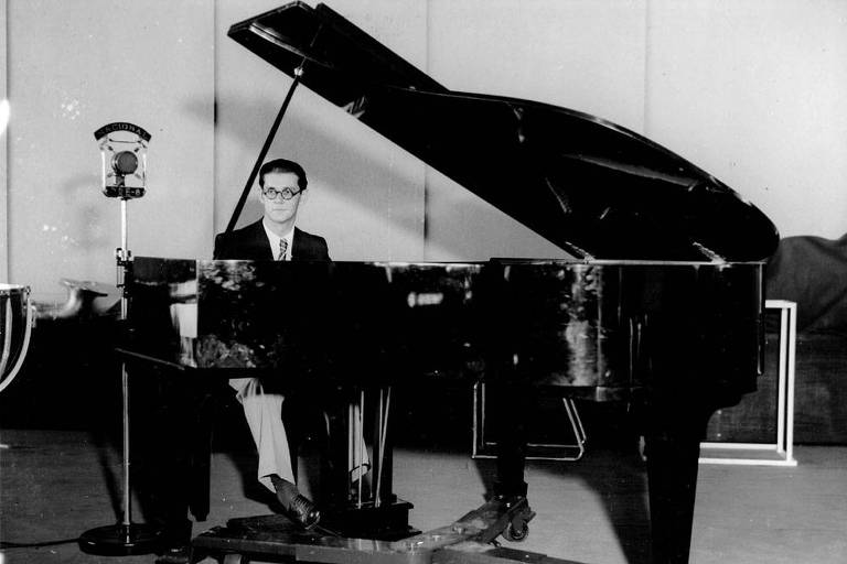 Em foto preto e branco Radamés Gnattali aperece sentado diante de um piano de meia calda com o tampo do instrumento aberto na Radio Nacional