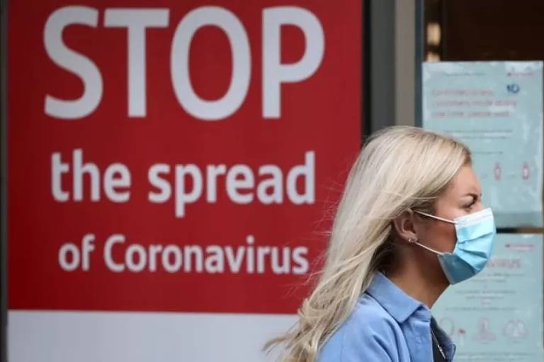 Mulher andando de máscara em frente a uma placa onde se lê "pare a transmissão do coronavírus"