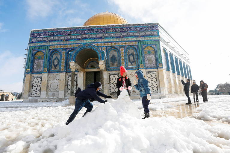 Crianças brincam com neve frente a construção religiosa histórica, em azul e branco, com domo dourado
