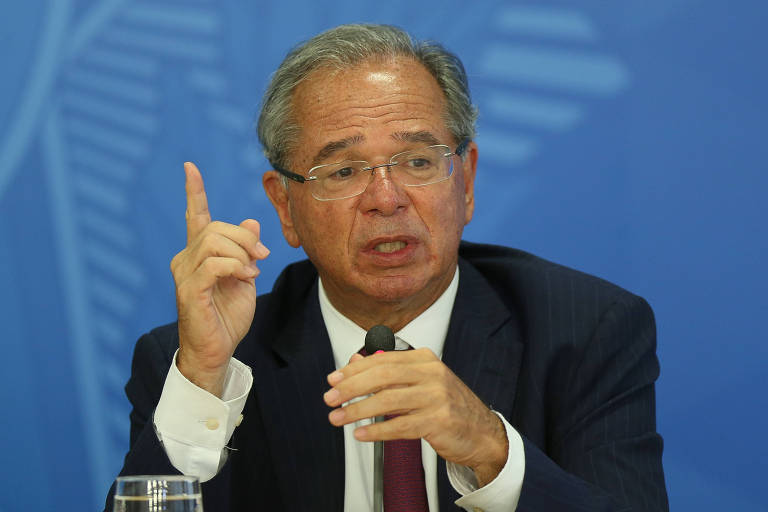 Paulo Guedes, de terno e gravata pretos e camisa branca, usando óculos, aponta para cima com o dedo indicador direito, em frente a um fundo azul
