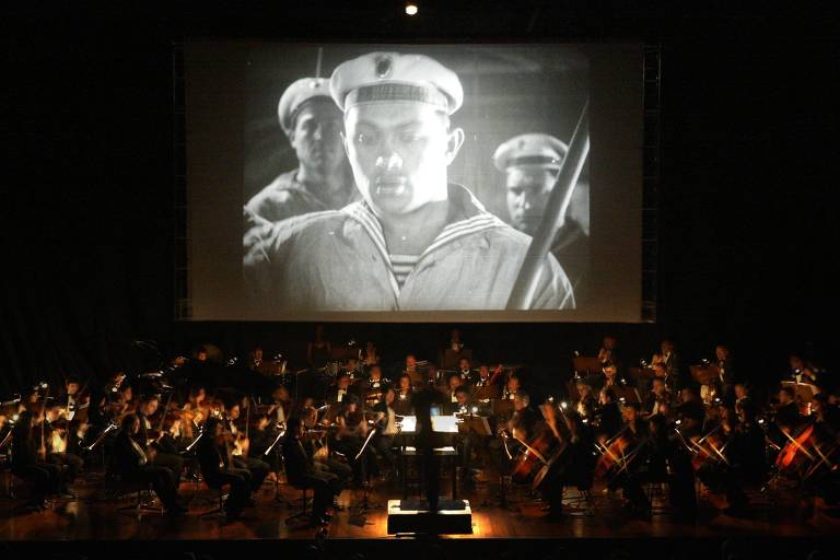 Em cima, um telão exibe cenas de marinheiros do filme 'O Encouraçado Potemkin', enquanto, à frente, é possível ver uma orquestra que se apresenta