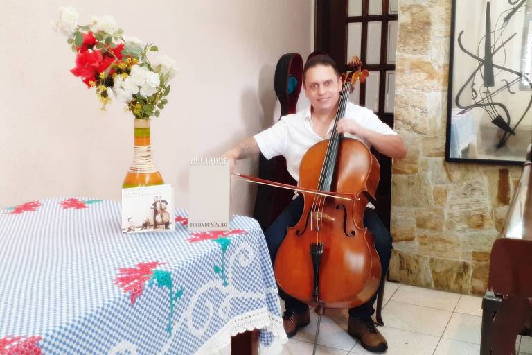 Em foto colorida, o violoncelista Mauro Brucoli aparece sentado na sala de sua casa tocando violoncelo