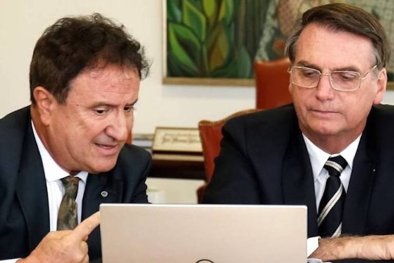 À esquerda, Evaristo de Miranda aponta o dedo indicador na tela de um computador à sua frente. Sentado ao seu lado está o presidente da República, Jair Bolsonaro, de óculos e braços cruzados. Ambos usam terno.