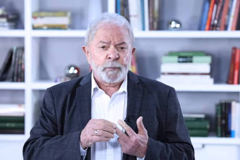 O ex-presidente Lula de camisa social branca com botão da gola aberto, paletó escuro; ao fundo, estante com livros e outros objetos