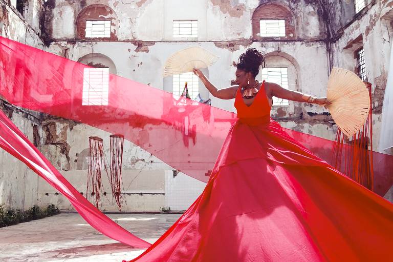 A cantora Margareth Menezes, uma mulher negra, está usando um vestido vermelho que tem um véu voando e segura um leque de palha na mão esquerda. Ela está em um casarão abandonado