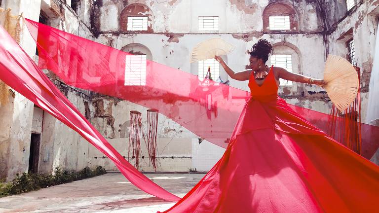 A cantora Margareth Menezes, uma mulher negra, está usando um vestido vermelho que tem um véu voando e segura um leque de palha na mão esquerda. Ela está em um casarão abandonado