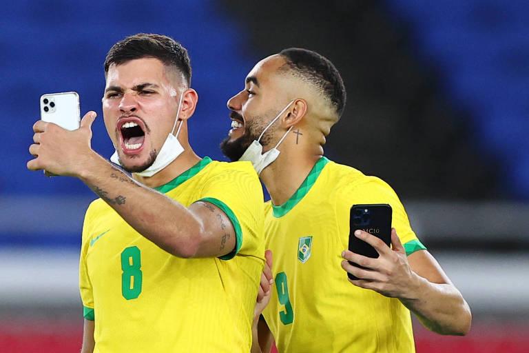 Ambos segurando celulares, Bruno Guimarães, que faz uma careta ao olhar para o aparelho, e Matheus Cunha celebram a conquista do ouro olímpico no futebol nas Olimpíadas de Tóquio pela seleção brasileira; os dois usam camisa amarela