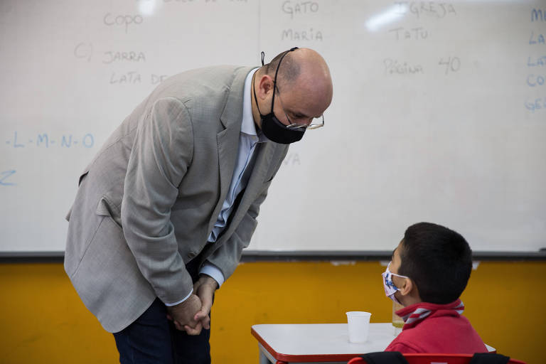 O secretário estadual da Educação de SP, Rossieli Soares, acompanha volta às aulas em novembro de 2021, abaixando-se para falar com aluno sentado em carteira escolar