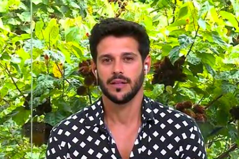 Com camisa preta de bolinhas brancas, Rodrigo dá entrevista sentado, com ambiente de flores verdes ao fundo 