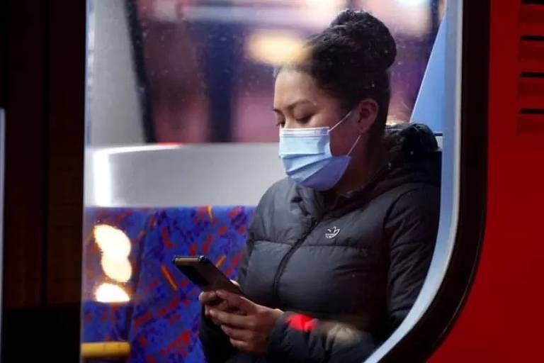 Imagem em primeiro plano mostra uma mulher negra de máscara segurando e olhando para a tela de um celular. Ela está sentada no banco de um transporte público.