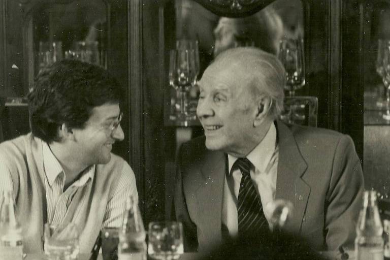 Dois homens brancos sentados se olham. Do lado esquerdo o jornalista Augusto Massi sorri para o argentino Jorge Luis Borges. Eles estão numa entrevista coletiva no hotel Maksud Plaza