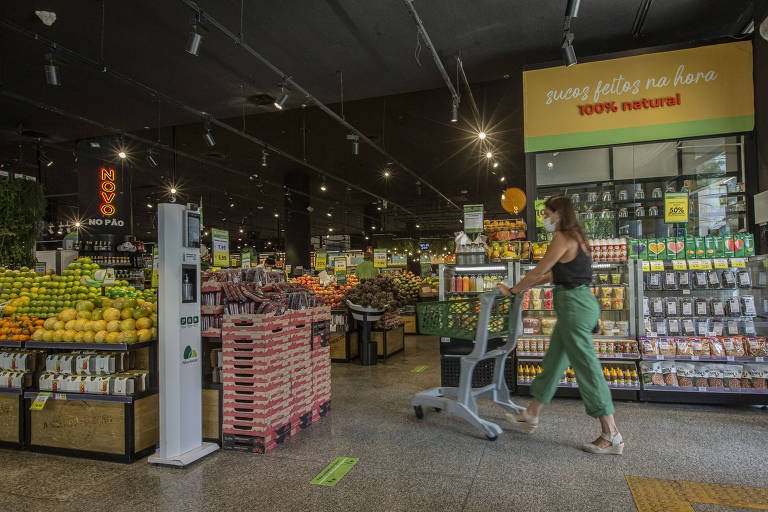 Mulher de blusa preta e calça verde empurra carrinho de compras em área de hortifruti de supermercado