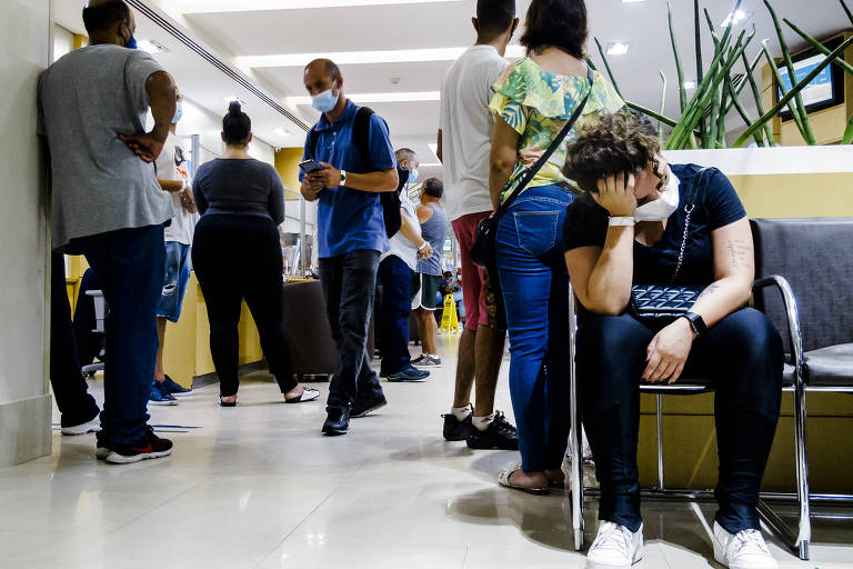 pessoas aglomeradas em uma recepção de hospital; mulher sentada, com mão na cabeça, em atitude de espera, usa máscara