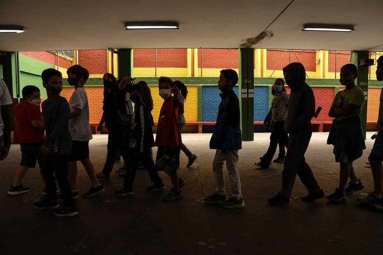 Várias crianças andando no interior de uma escola, vestidas com roupas coloridas e usando máscaras de proteção contra o coronavírus. Ao fundo, vê-se parte do prédio da escola, com paredes pintadas de vermelho, azul, verde e amarelo