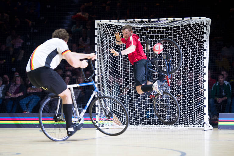 dois ciclistas, uma bola e as traves de um gol.