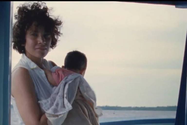 Uma mulher branca de cabelos cacheados escuros e curtos está em pé numa janela segurando um bebê