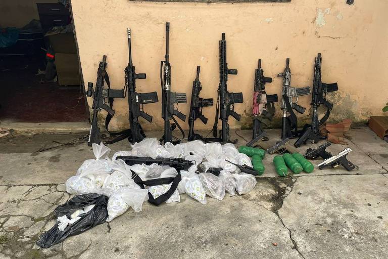 Imagem mostra 8 fuzis enconstados em parede, cinco pistolas no chão e outros materias em sacos plásticos