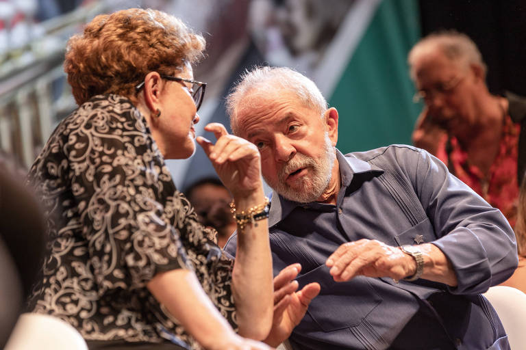 Dilma de óculos cochicha com Lula, sentados, em palco de evento