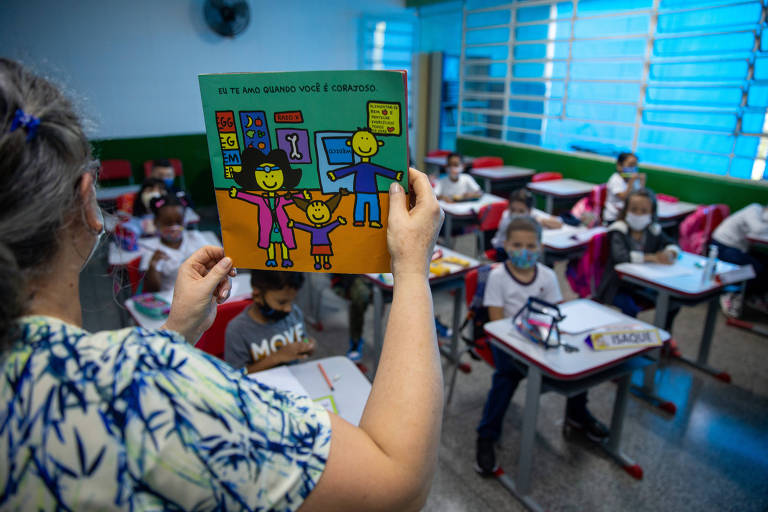 Professora mostra livro infantil, com capa colorida, para alunos que a observam sentados em suas carteiras