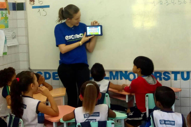 Crianças são vistas de costas em uma sala de aula; diante delas uma professora segura um celular
