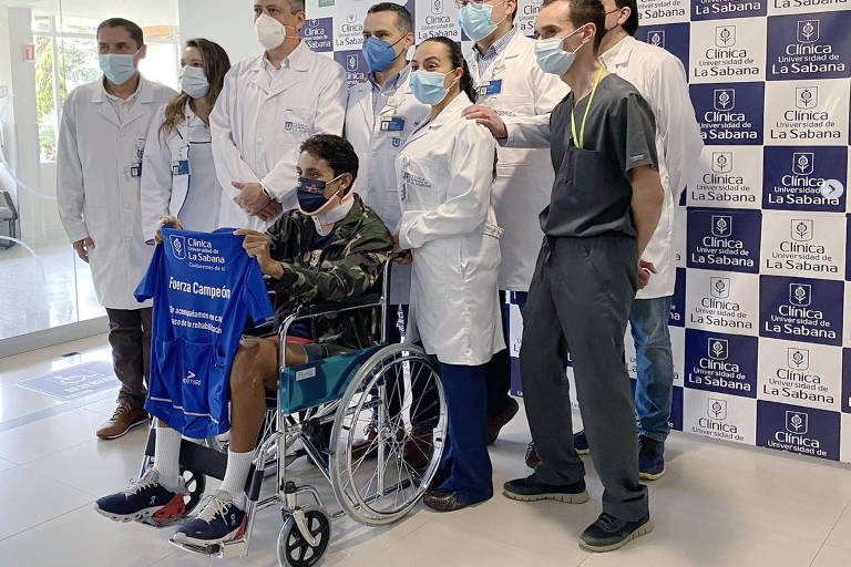Cercado por médicos, o atleta segura uma camiseta, sentado em cadeira de rodas