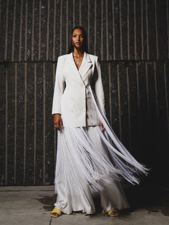 A modelo Lais Ribeiro, mulher negra, está em pé com uma saia branca esvocaçante e um paletó branco. Ela usa o cabelo preso.