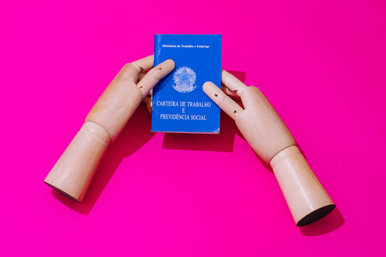 Foto mostra mãos de madeira segurando carteira de trabalho, em fundo rosa