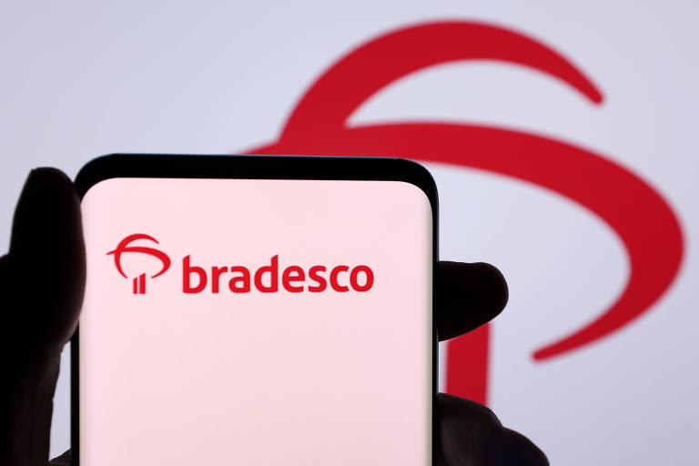 Imagem mostra uma mão segurando um celular, no qual é possível ver o logo do banco Bradesco, em vermelho. Atrás, há um painel com a logo, que é formada por dois traços na vertical e dois riscos curvados que se cruzam, em vermelho.