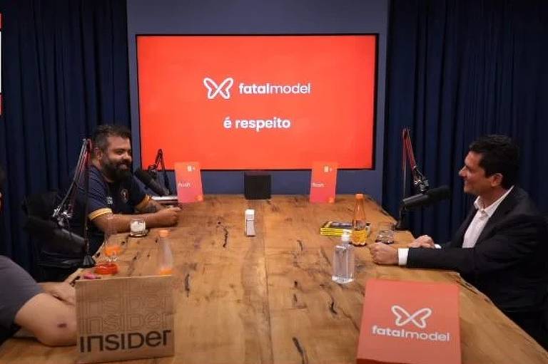 Sergio Moro e Monark estão sentados em uma mesa com microfones em frente a eles