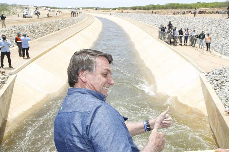 Em frente a um trecho do rio São Francisco transposto, Bolsonaro, de camisa azul, faz o sinal de joia com as duas mãos e sorri
