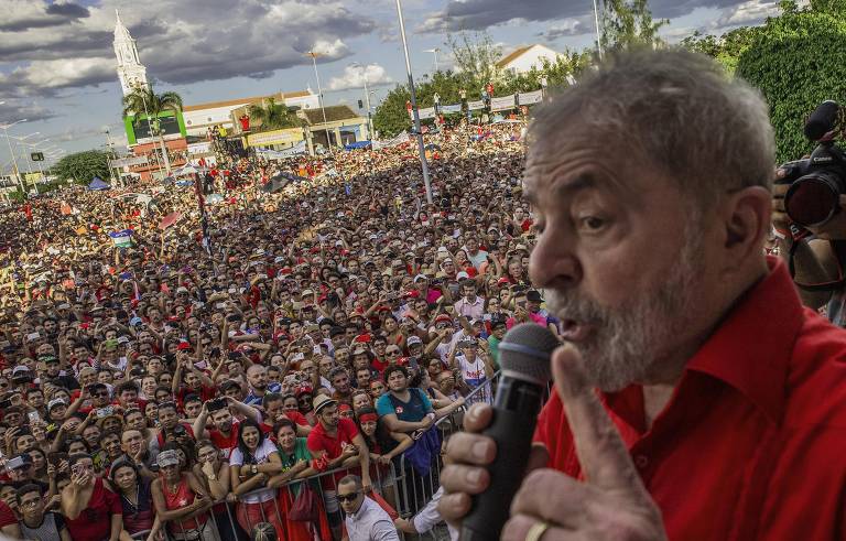 Lula, de camisa vermelha e com microfone na mão, está à direita da foto; ao fundo, no chão, multidão