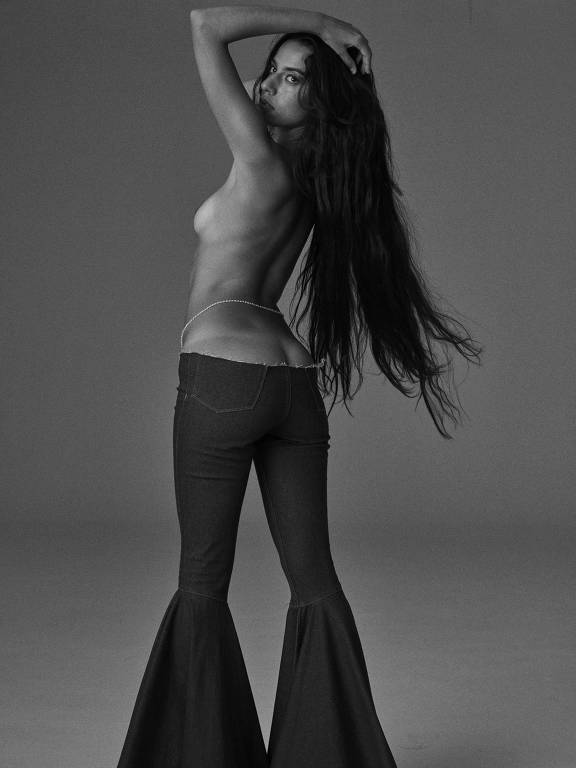 A cantora Marina Sena, mulher branca de cabelos negros com comprimento até o final das costas, está em pé, de perfil, usando apenas calça jeans, com parte do bumbum e do seio esquerdo à mostra
